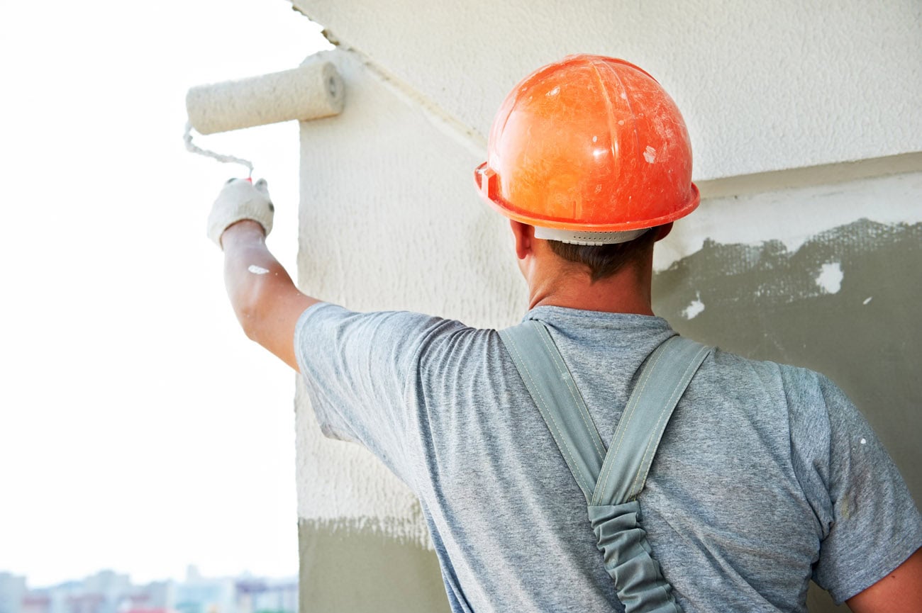Imbianchino al lavoro su parete esterna Imbianchino con casco arancione che dipinge parete esterna con rullo.
